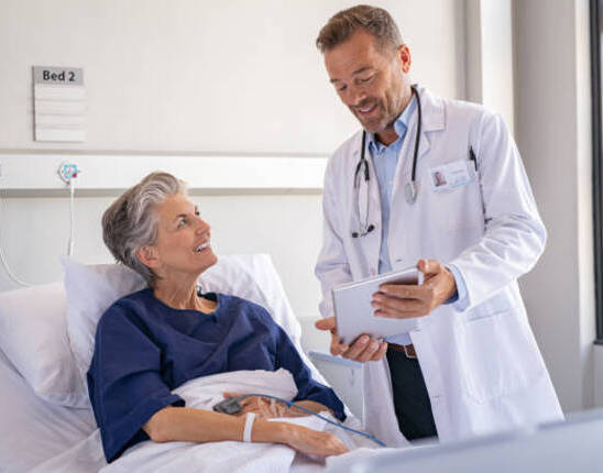 Smiling senior woman sitting on hospital bed listening to mature surgeon at private clinic. Confident specialist in labcoat holding digital tablet and talking to old patient. Happy professional friendly doctor meeting elderly patient on a hospital bed and explaining health results.